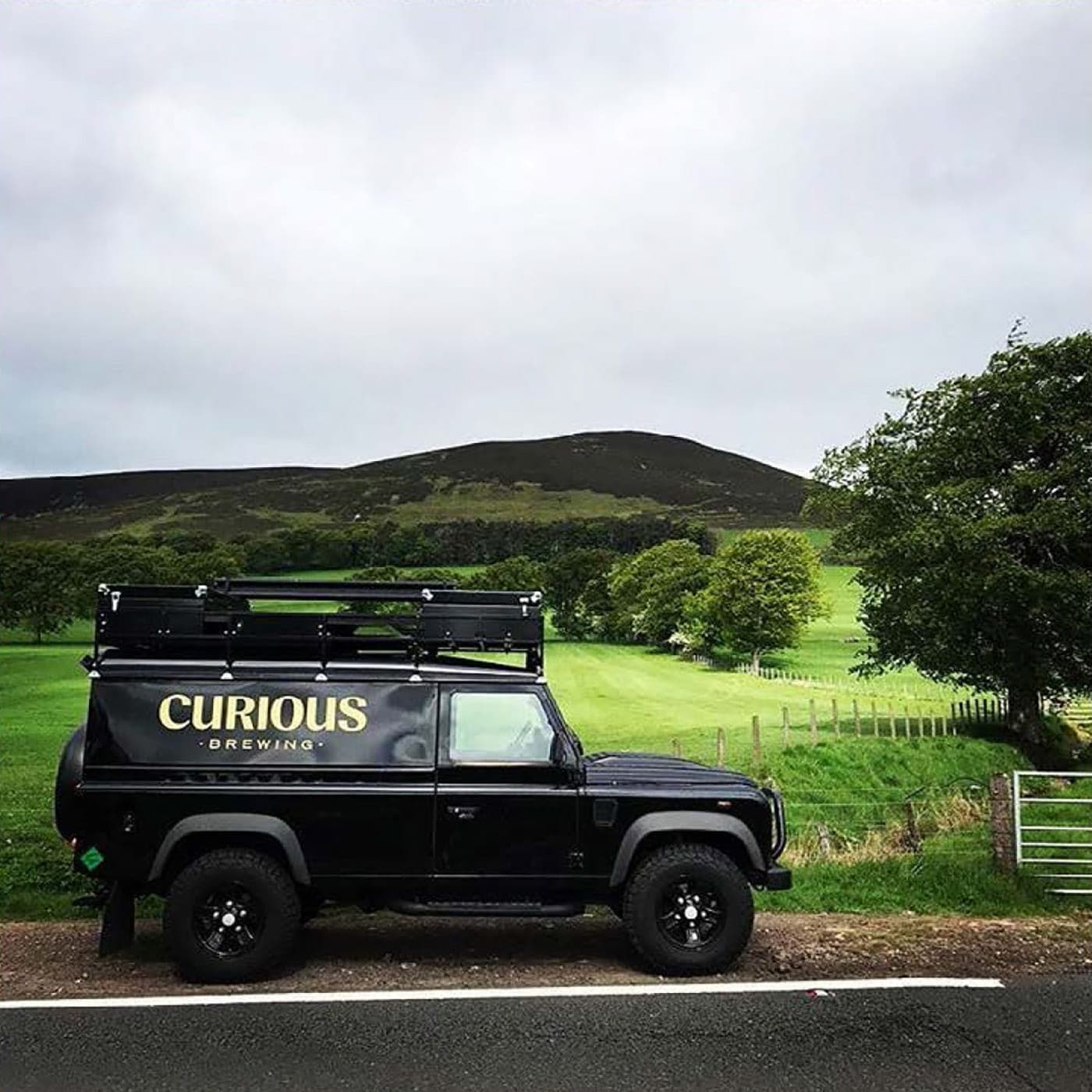 The black Curious Brewing branded Land Rover parked in green countryside