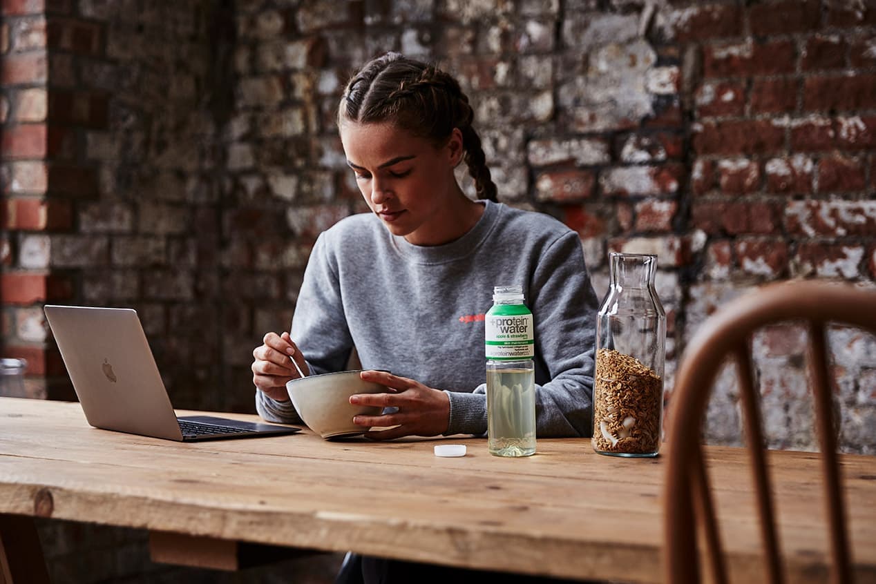 A woman having breakfast at a laptop on a wooden table, with a bottle of Protein Water