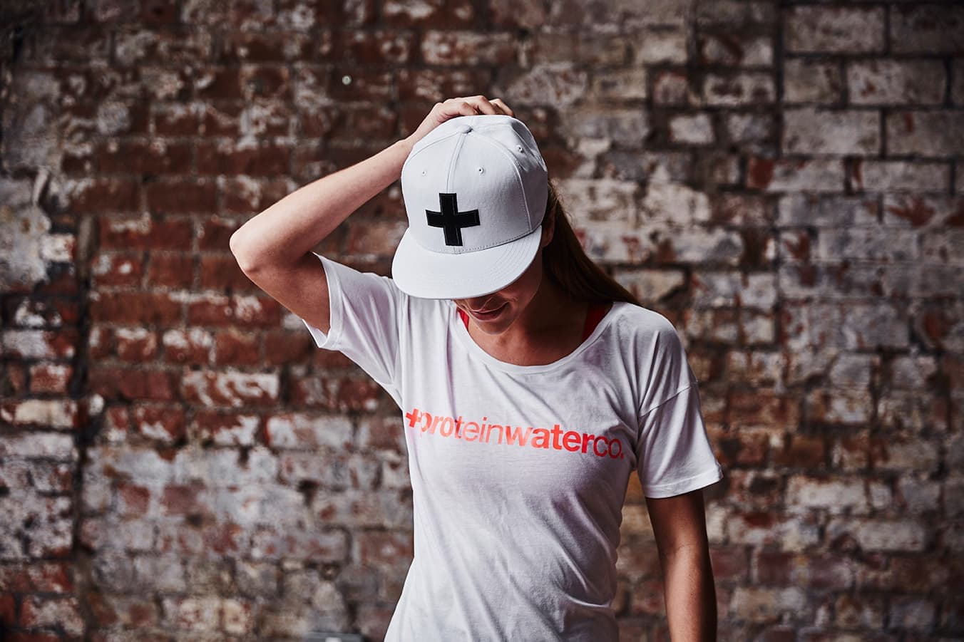 A woman in a Protein Water Co. T-shirt and a white cap with the brand's plus symbol