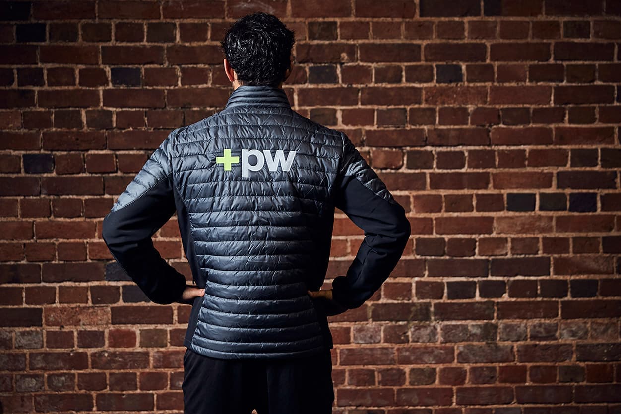 A man in a +pw branded padded jacket, seen from behind against a brick wall