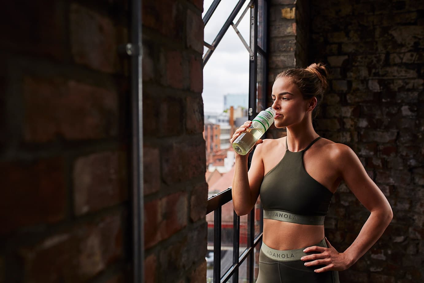 A woman in sports kit drinking Protein Water by an open window in a warehouse studio