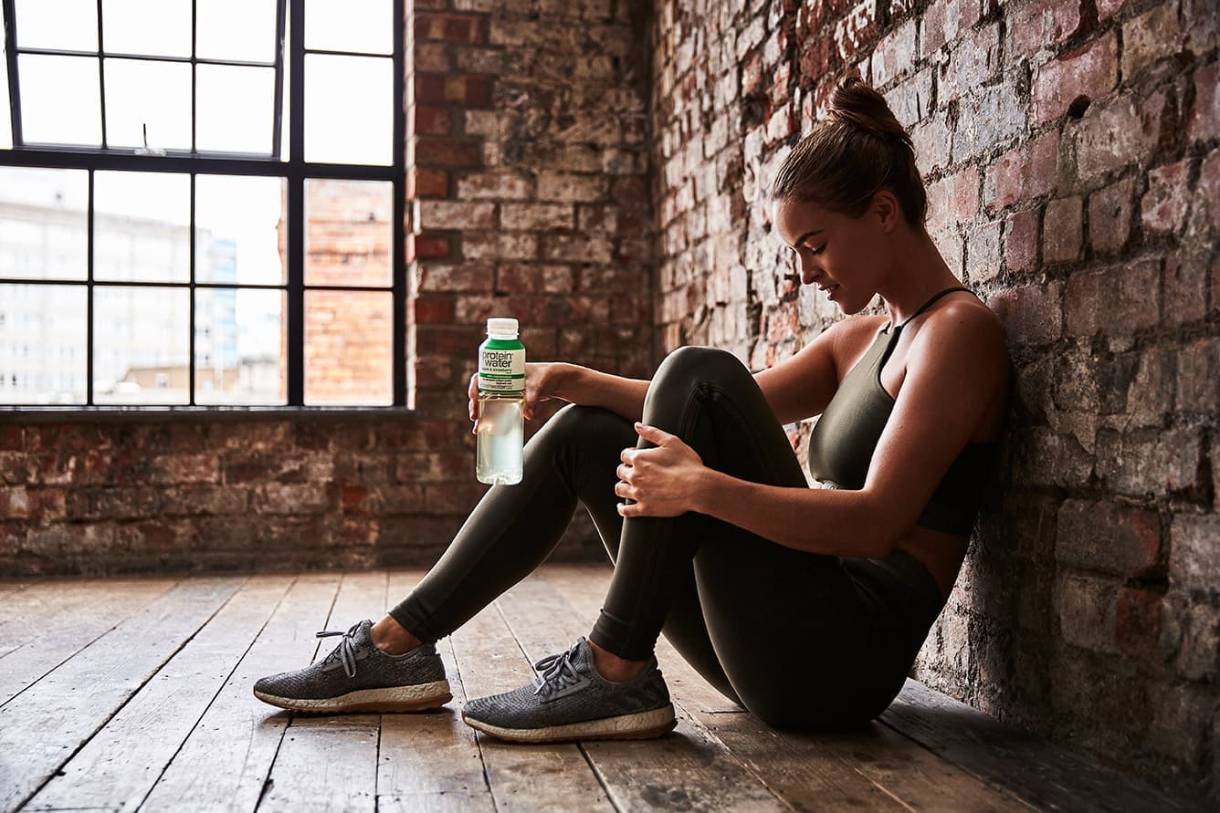 A woman in sports kit resting against a brick wall with a bottle of Protein Water
