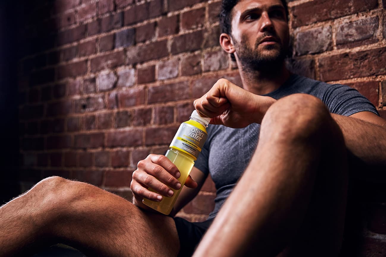A man sitting on the gym floor between sets, holding a bottle of lemon Protein Water