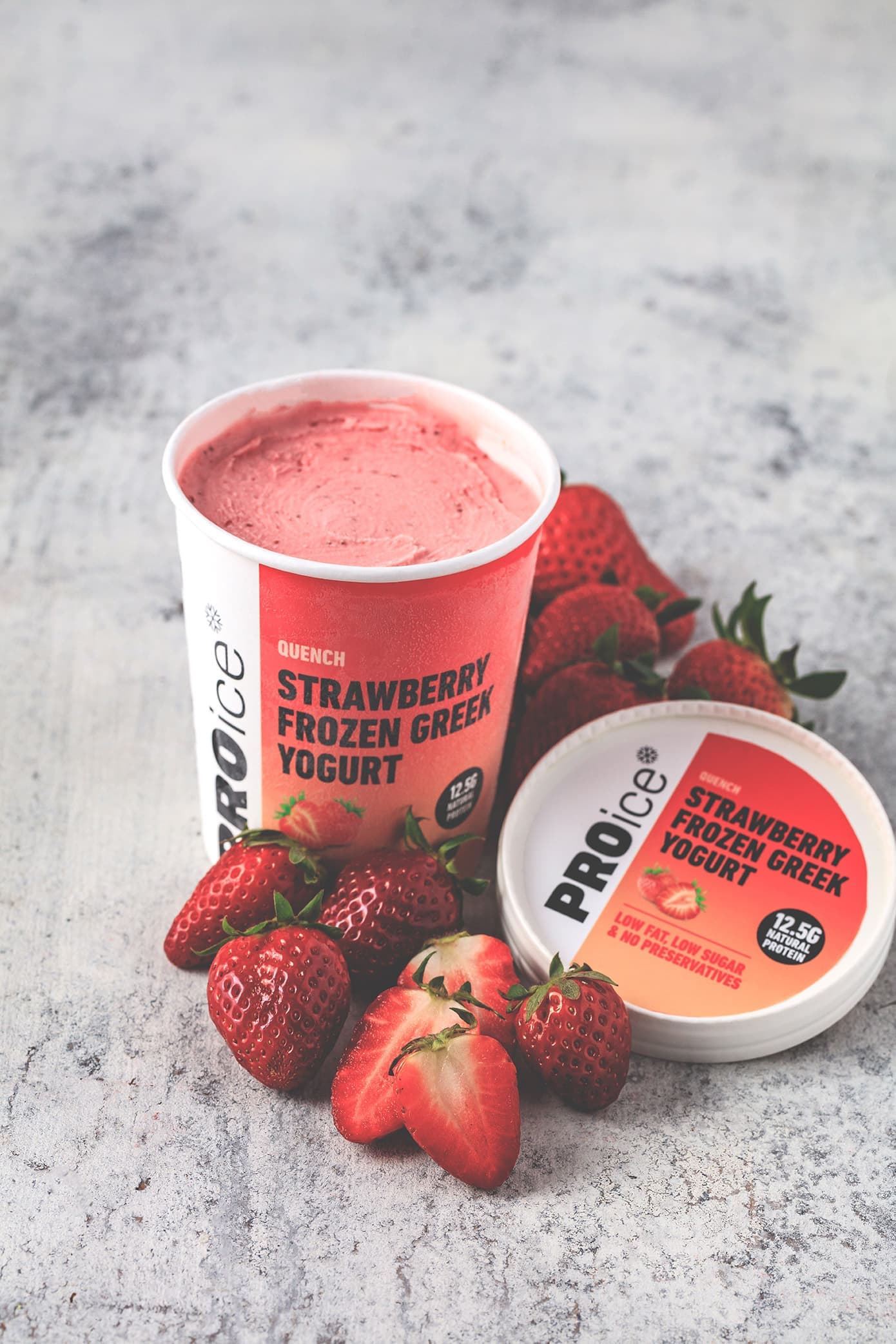 A tub of PROice Strawberry Frozen Greek Yogurt with fresh strawberries on a grey stone surface