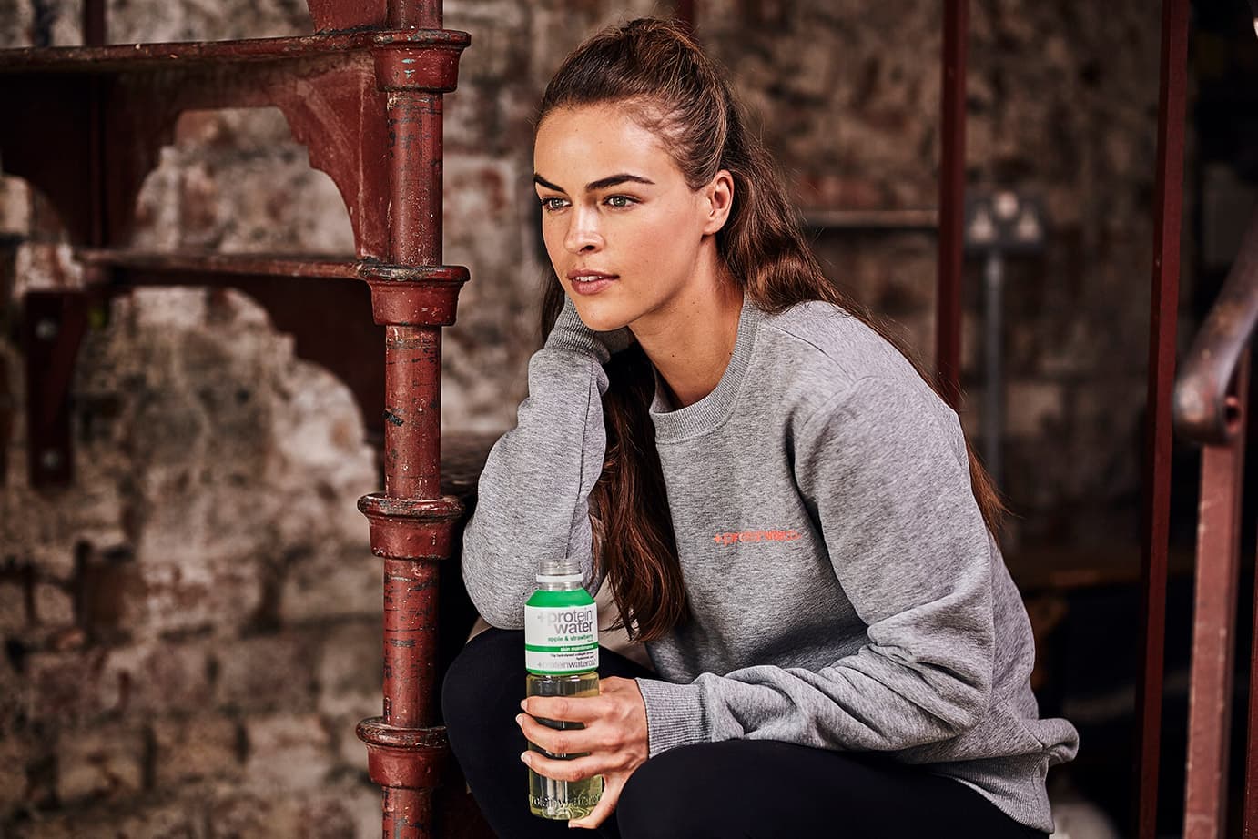 A woman in a grey Protein Water Co. sweatshirt sitting on an industrial staircase, holding a bottle of Protein Water