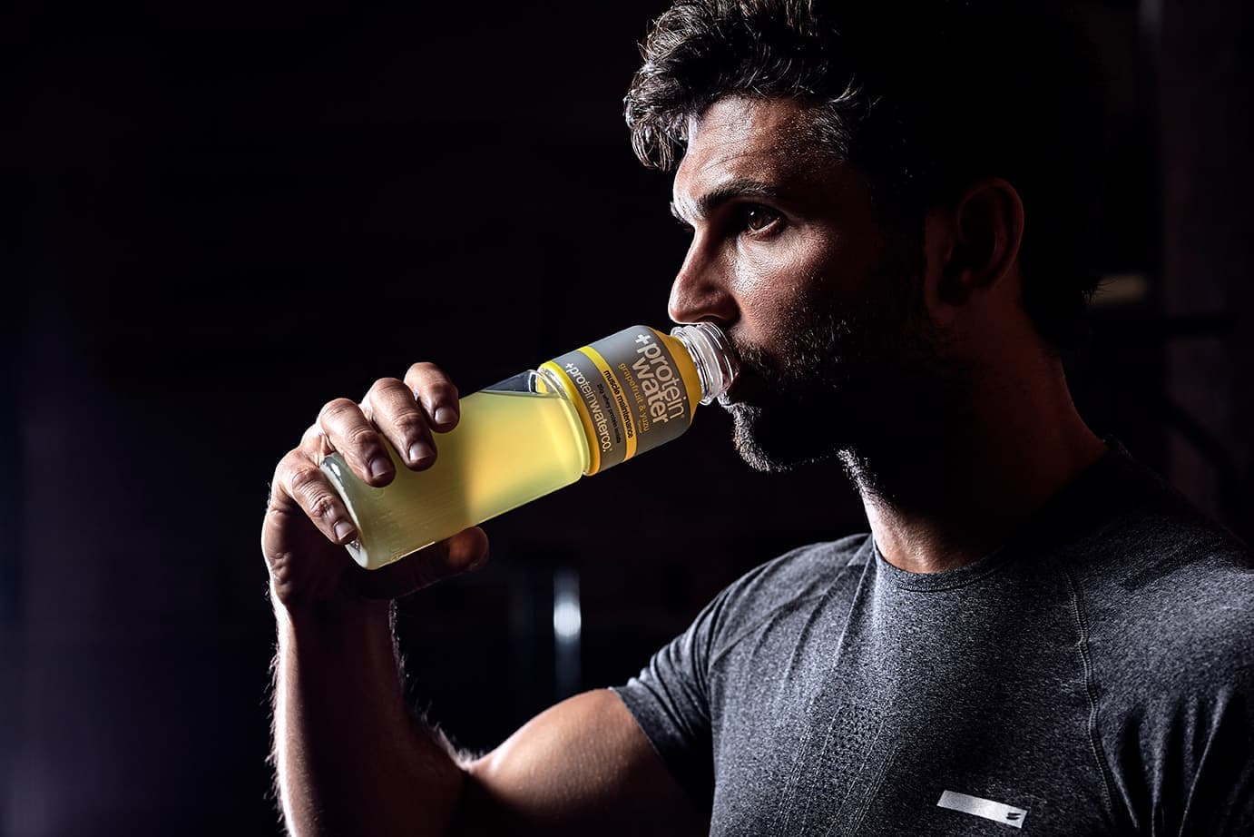 A man drinking from a bottle of lemon Protein Water in low light