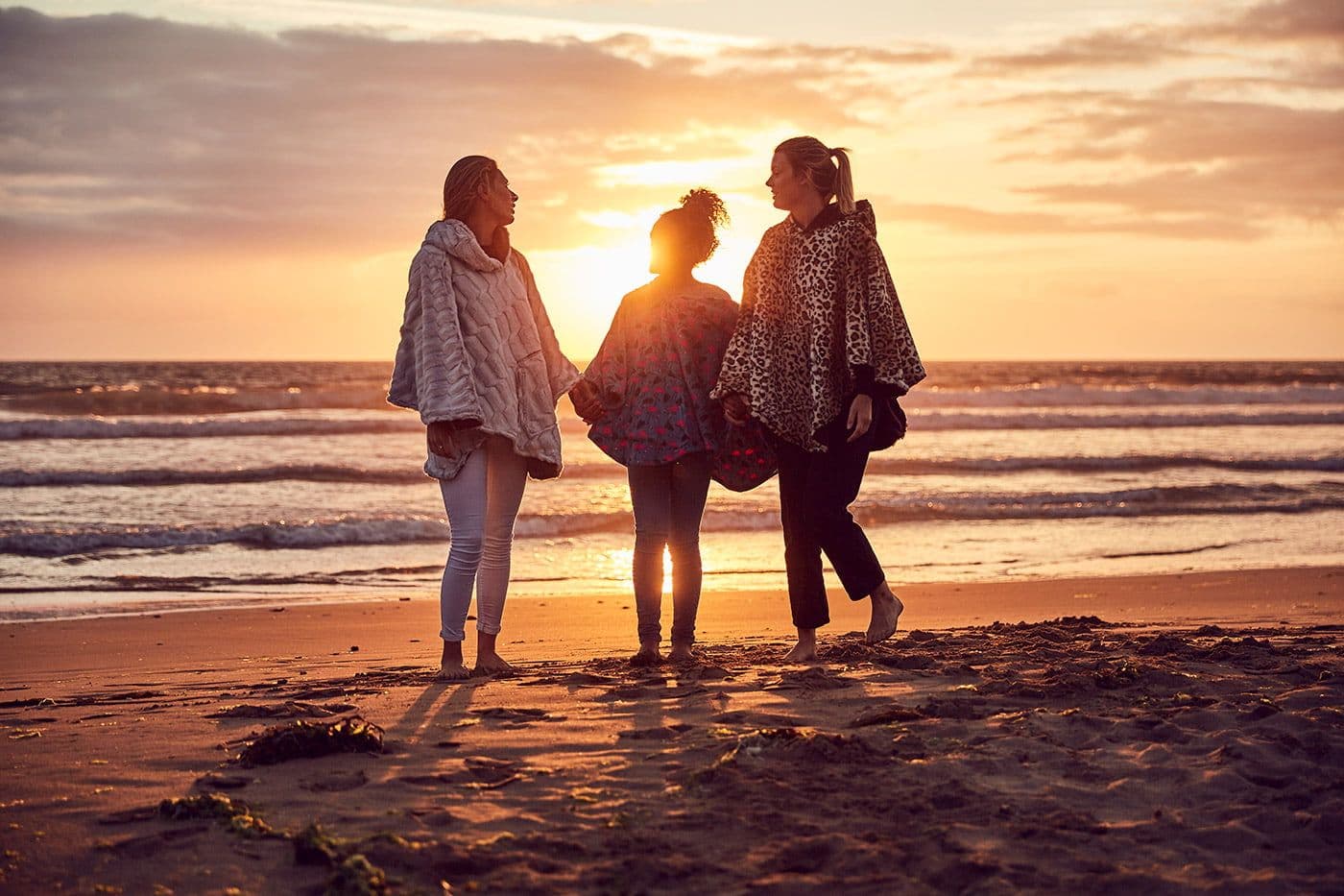 Three women in Ponchella ponchos on a beach at sunset