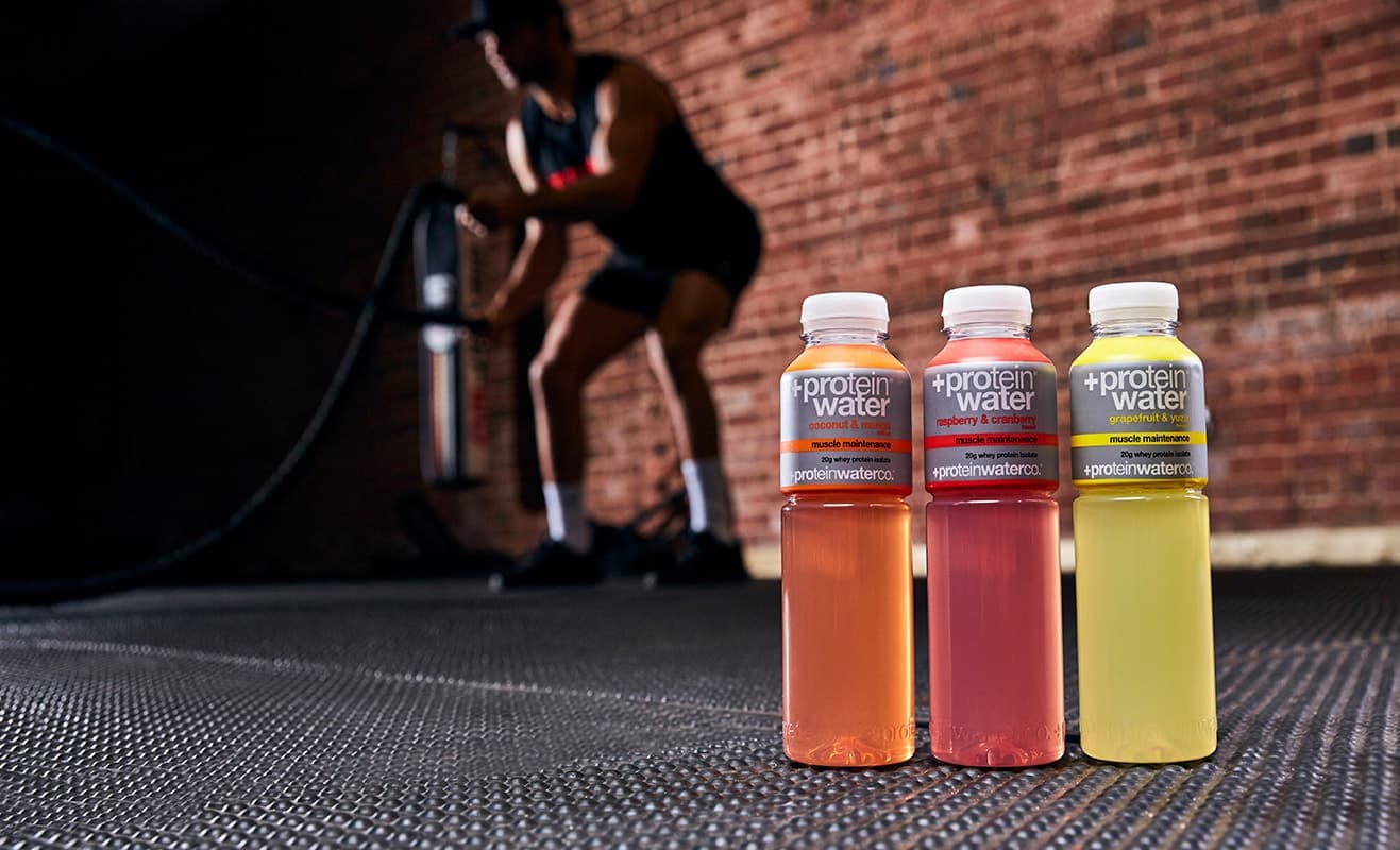 Three bottles of Protein Water on a gym floor, with a man training with battle ropes behind