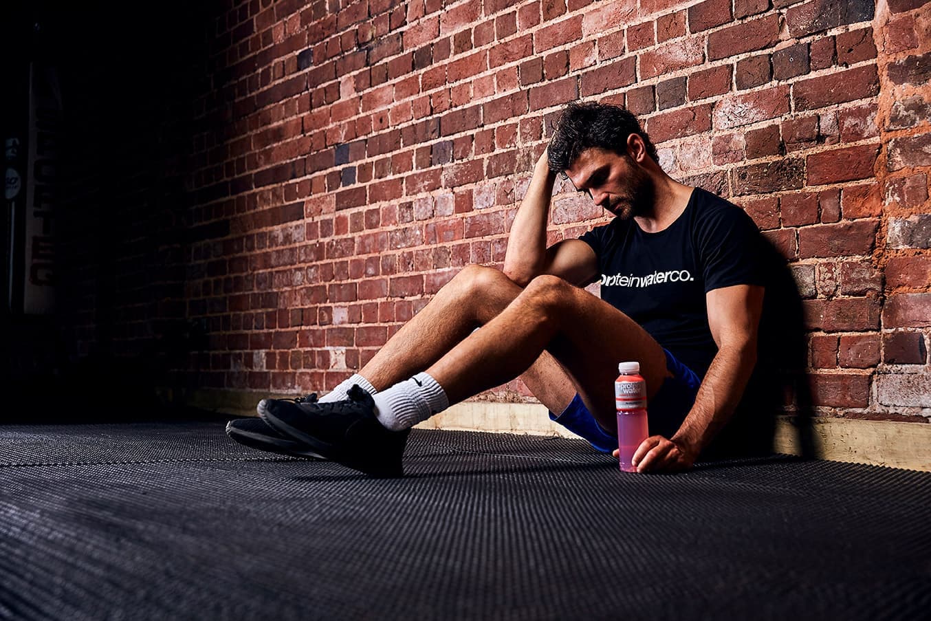 A man resting against a brick wall after a workout, with a bottle of Protein Water beside him