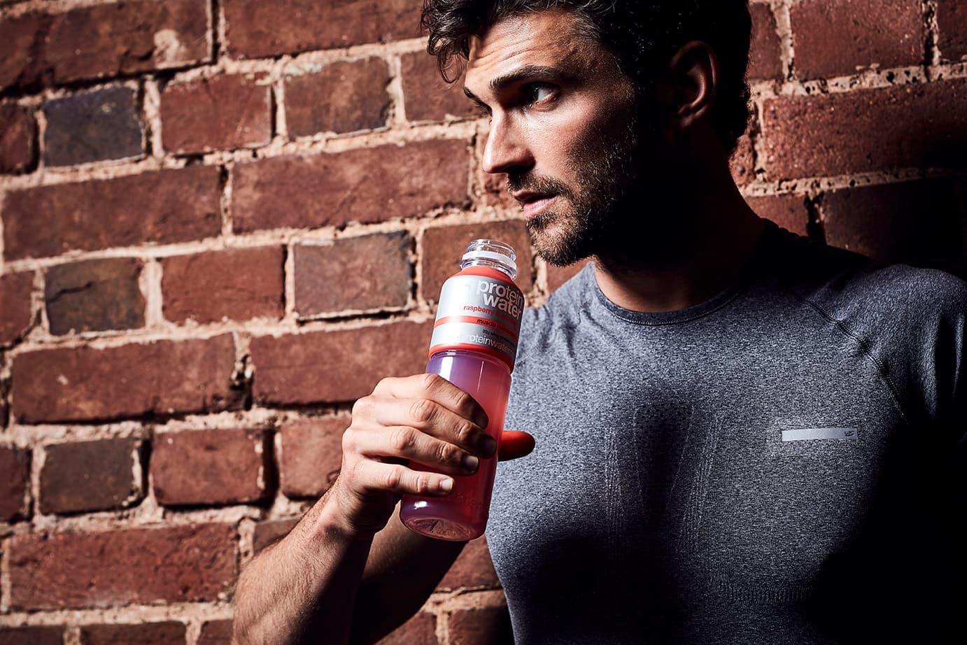 A man in a grey training top holding a bottle of raspberry Protein Water against a brick wall